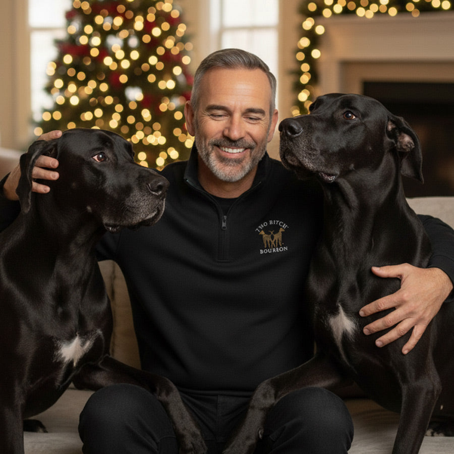 Man sitting with two black dogs in a room decorated for Christmas. The man is wearing a two Bitch Bourbon embroidered Zip Pullover Sweatshirt.