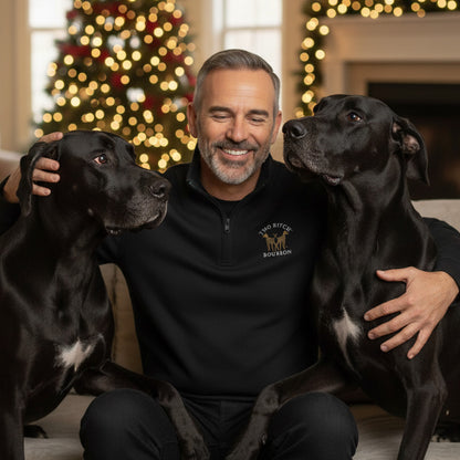 Man sitting with two black dogs in a room decorated for Christmas. The man is wearing a two Bitch Bourbon embroidered Zip Pullover Sweatshirt.