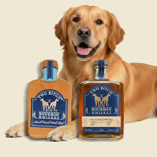 Dog with a bottle of 'Two Bitch Bourbon Whiskey' on a beige background